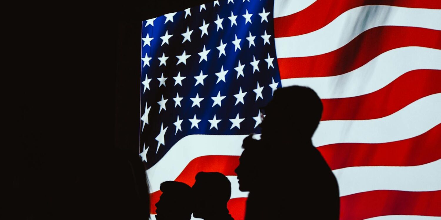 Silhouetted figures in front of a bright, wavy American flag create a patriotic scene.